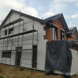 Dom w trakcie ocieplania: białe płyty izolacyjne na ścianach, rusztowanie, częściowo odkryta cegła, czarna folia ochronna, widoczny dach z czarną dachówką.
