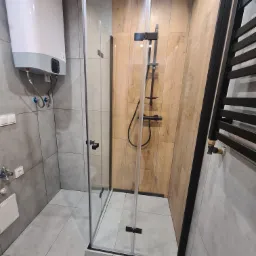 Nowoczesna łazienka z kabiną prysznicową typu walk-in, drewnopodobnymi panelami na ścianie i grzejnikiem drabinkowym w kolorze czarnym.