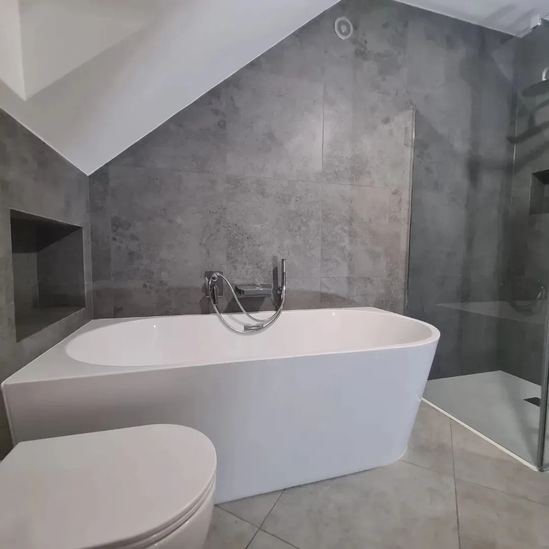 Nowoczesna łazienka w odcieniach szarości z białą, prostokątną wanną i kabiną prysznicową typu walk-in, wykończona płytkami imitującymi beton i z wnęką w ścianie.