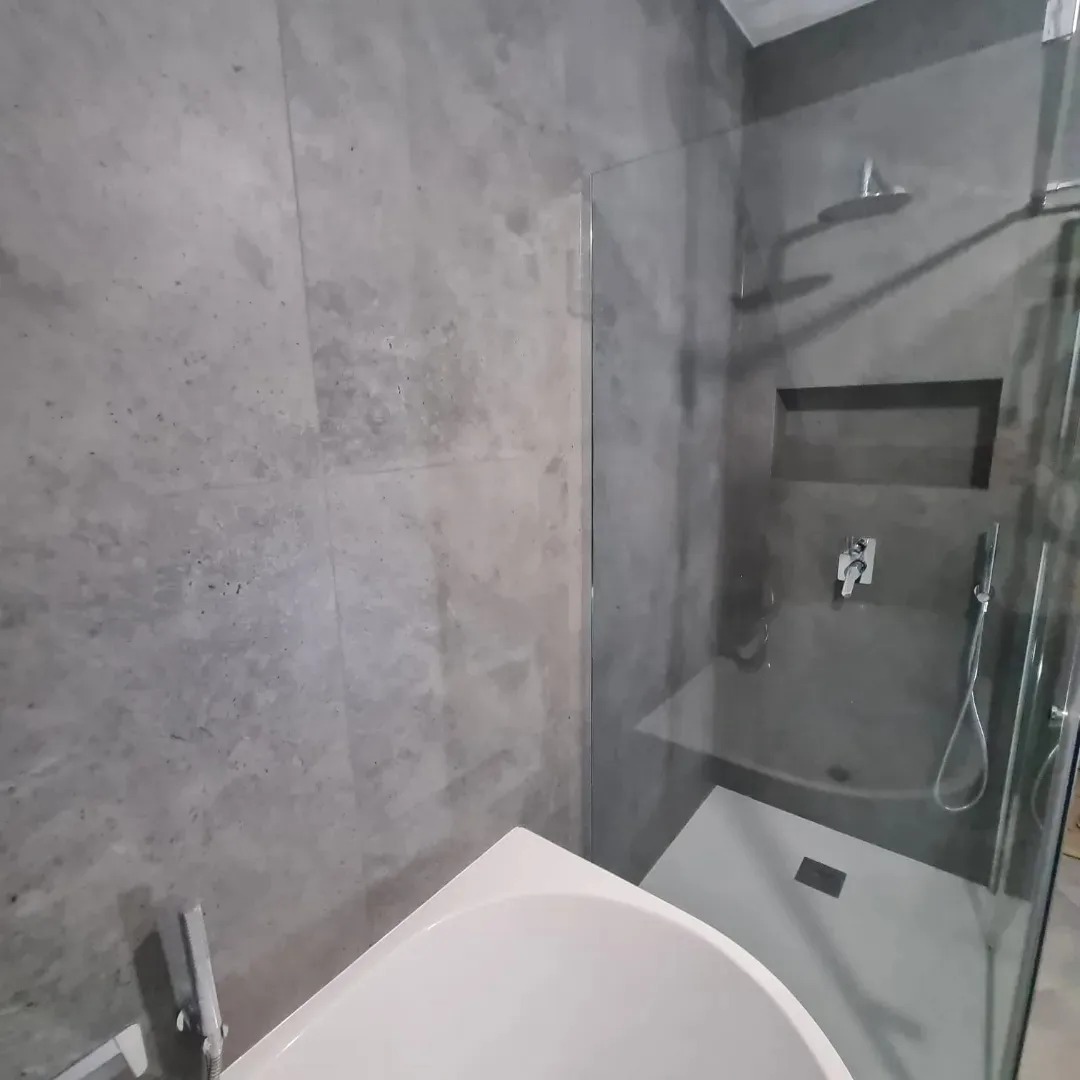 Nowoczesna łazienka z szarymi betonowymi ścianami, kabiną prysznicową typu walk-in z deszczownicą i wnęką na kosmetyki, oraz fragmentem białej wanny.