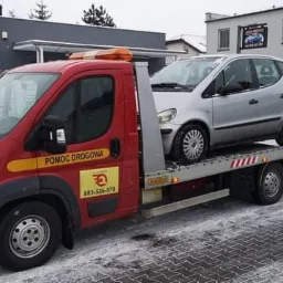Czerwona laweta z napisem 'Pomoc Drogowa' i numerem telefonu przewozi srebrny samochód osobowy na tle zaśnieżonego krajobrazu miejskiego.