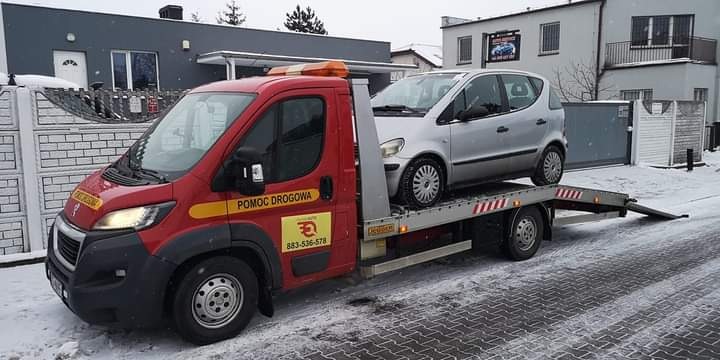 Czerwona laweta z napisem 'Pomoc Drogowa' i numerem telefonu przewozi srebrny samochód osobowy na tle zaśnieżonego krajobrazu miejskiego.