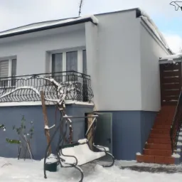 Ośnieżony dwukondygnacyjny dom z balkonem i schodami zewnętrznymi, pomalowany na biało z szarym cokołem, huśtawka ogrodowa pokryta śniegiem, logo firmy Bergman w prawym górnym rogu.