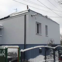 Ocieplony dom jednorodzinny w kolorze szarym z niebieskim cokołem, częściowo zasłonięty przez ogrodzenie i śnieg, z widocznym logo firmy Bergman w prawym górnym rogu.