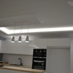 Nowoczesna, biała kuchnia z drewnianym blatem, oświetleniem podszafkowym LED i trzema wiszącymi lampami w kształcie kropli, czarny piekarnik w zabudowie, logo firmy Bergman w prawym g&oacute;rnym rogu.