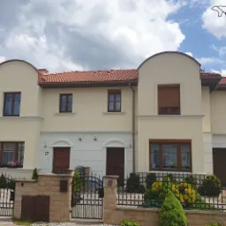 Dwukondygnacyjny dom bliźniak z elewacją w odcieniach beżu i brązu, z czerwonym dachem z dachówki, widoczny zza ozdobnego, czarnego ogrodzenia z kamiennymi słupkami i skrzynką na listy, w tle...