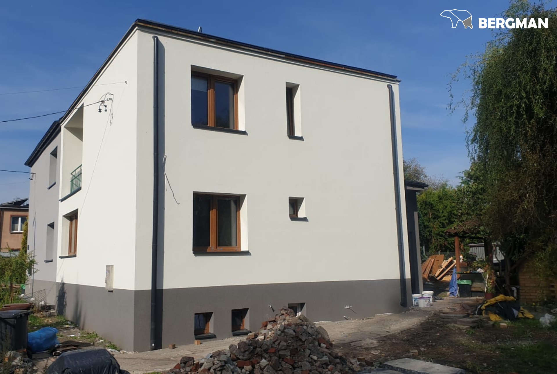 Dwukondygnacyjny budynek z jasną elewacją i ciemnoszarym cokołem, widoczne brązowe ramy okienne, na pierwszym planie sterta gruzu, w tle zieleń drzew i krzewów.