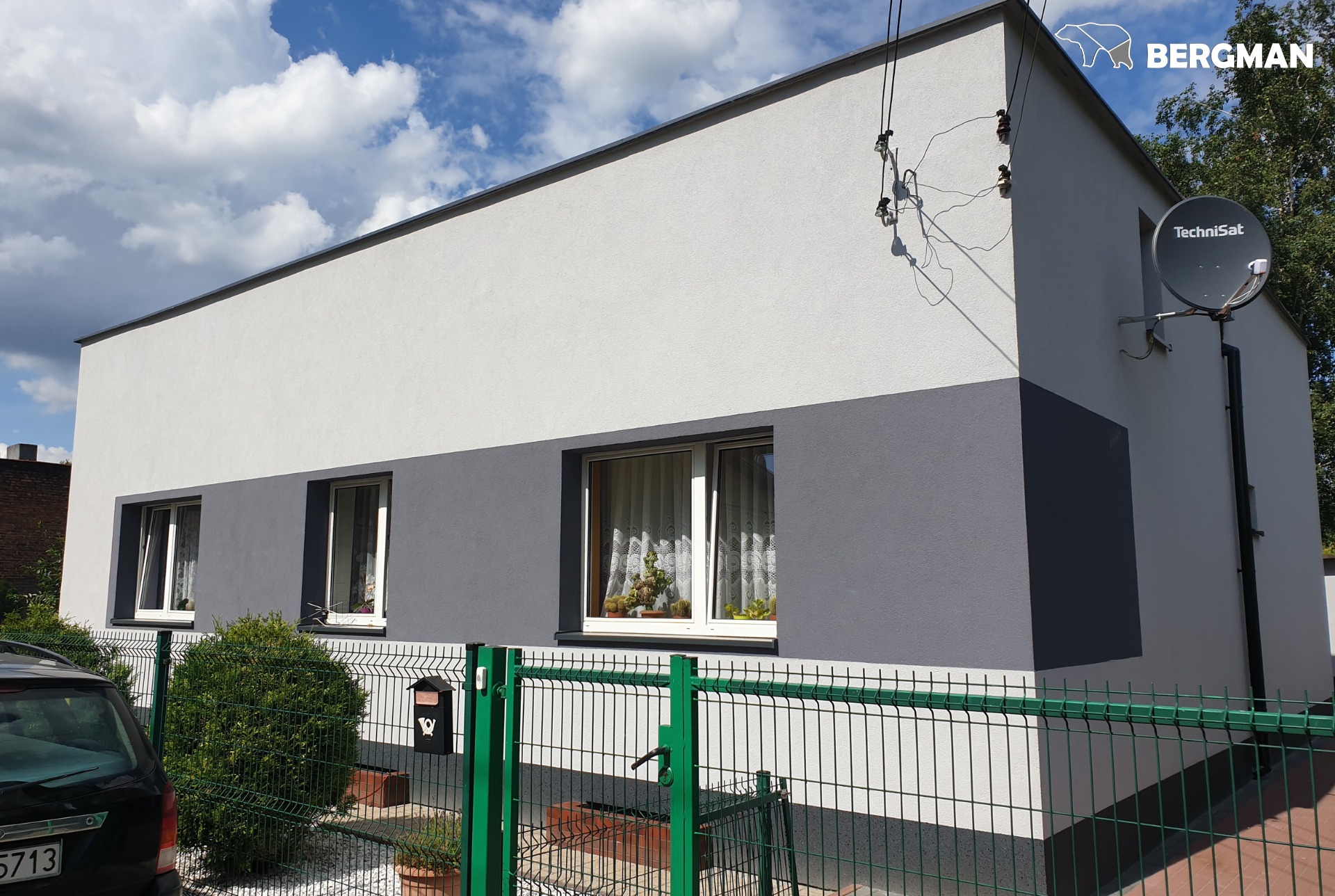 Dwukolorowa elewacja domu jednorodzinnego z szarą dolną częścią i białą górną, widoczna antena satelitarna, zielone ogrodzenie siatkowe i częściowo widoczny samochód na pierwszym planie.