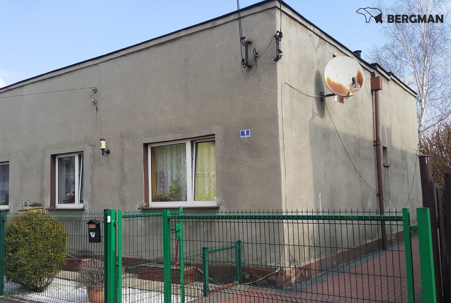 Szary, nieocieplony dom jednorodzinny z widoczną, rdzewiejącą anteną satelitarną, zielonym ogrodzeniem i numerem posesji '1 Marynarska'.