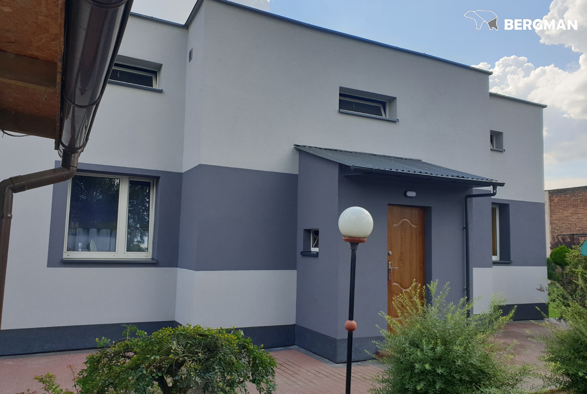 Dwukolorowa elewacja domu z drewnianymi drzwiami wejściowymi, widoczne rynny i lampa ogrodowa, logo firmy Bergman w prawym górnym rogu.
