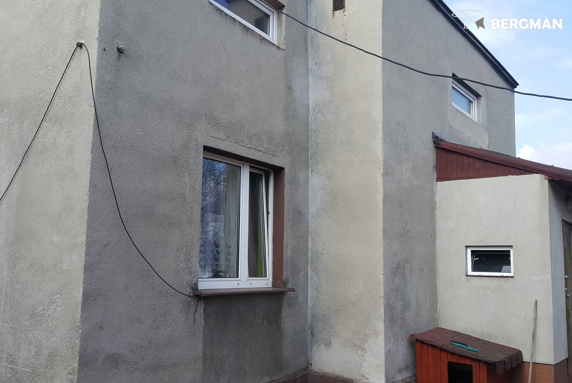 Szary, nierówny tynk na ścianie domu z widocznymi pęknięciami i śladami napraw, okno z białymi ramami i otwartym skrzydłem, oraz fragment dachu budki dla psa