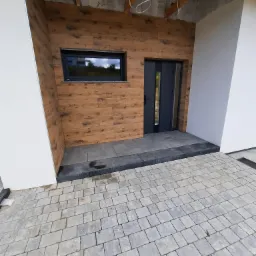 Wejście do domu z drewnianą okładziną elewacyjną, szarą stolarką okienną i drzwiową oraz wybrukowanym podjazdem z kostki w odcieniach szarości.