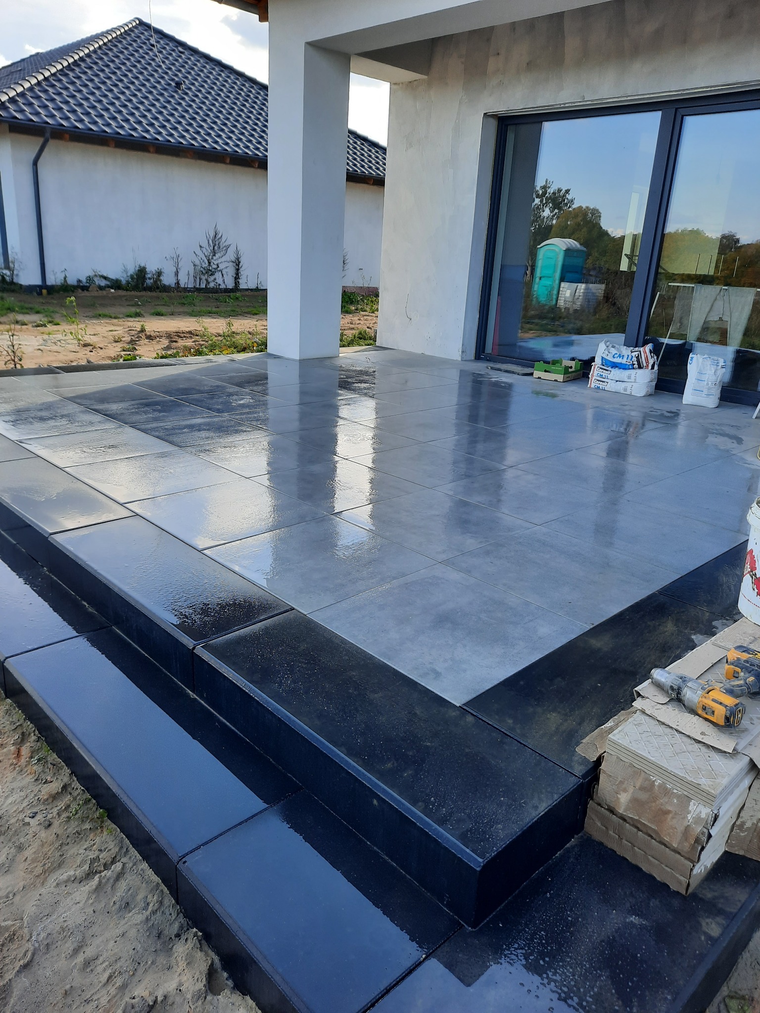 Świeżo ułożona, błyszcząca, szara płytka na zewnątrz budynku, z czarnymi obrzeżami schodów. Na płytkach widoczne odbicia nieba i budynku. W tle worki z zaprawą i narzędzia.