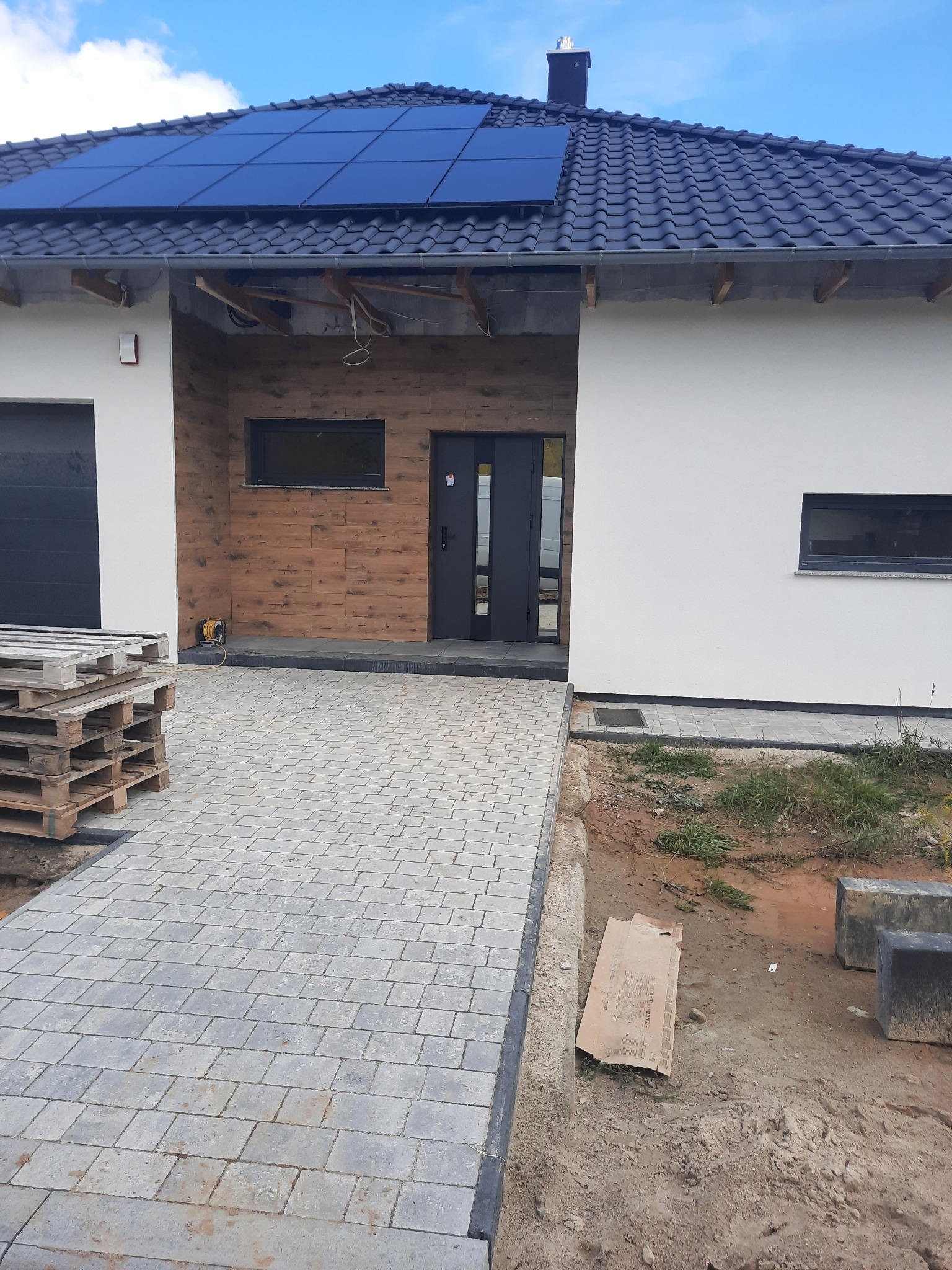 Nowoczesny dom jednorodzinny z panelami słonecznymi na dachu, wykończony drewnianą elewacją przy wejściu i brukowanym podjazdem, palety z materiałami budowlanymi obok garażu.