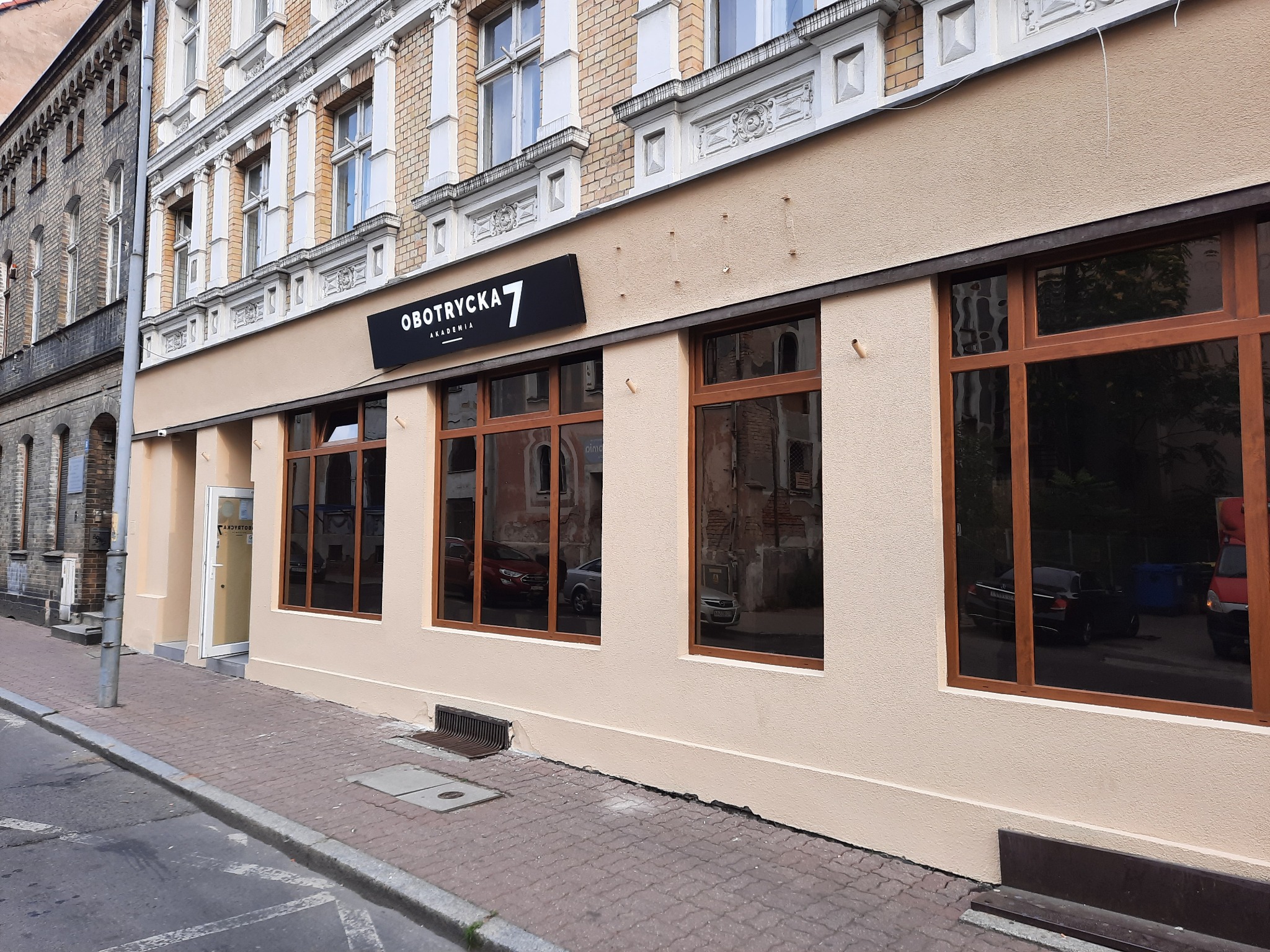 Odnowiona fasada budynku z beżową elewacją, ciemnobrązowymi ramami okiennymi i szyldem 'Obotrycka 7 Akademia' nad wejściem, widok z poziomu ulicy.