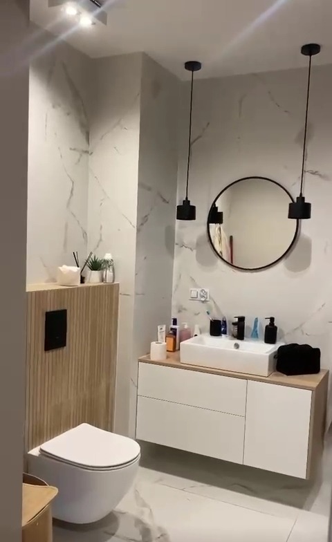 Nowoczesna łazienka z marmurowymi ścianami, wiszącą toaletą, szafką z umywalką i okrągłym lustrem, oświetlona dwoma czarnymi lampami wiszącymi.