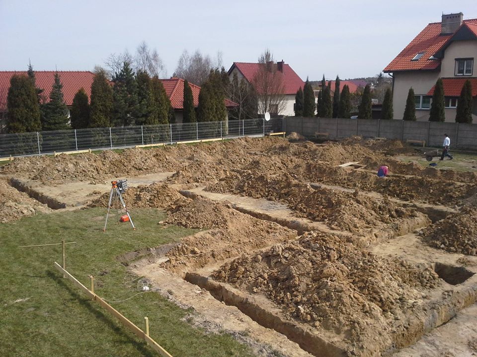 Wykonane wykopy pod fundamenty domu jednorodzinnego, widoczny niwelator na trójnogu, częściowo trawnik, w tle zabudowa jednorodzinna.