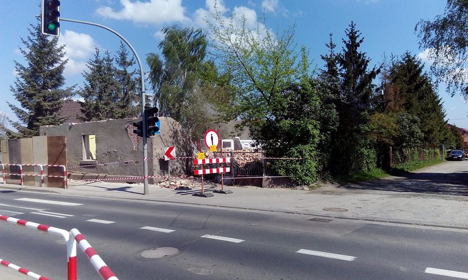 Częściowo zburzony budynek z odsłoniętą cegłą, zabezpieczony taśmą ostrzegawczą i znakami drogowymi na tle zieleni i błękitnego nieba z chmurami.