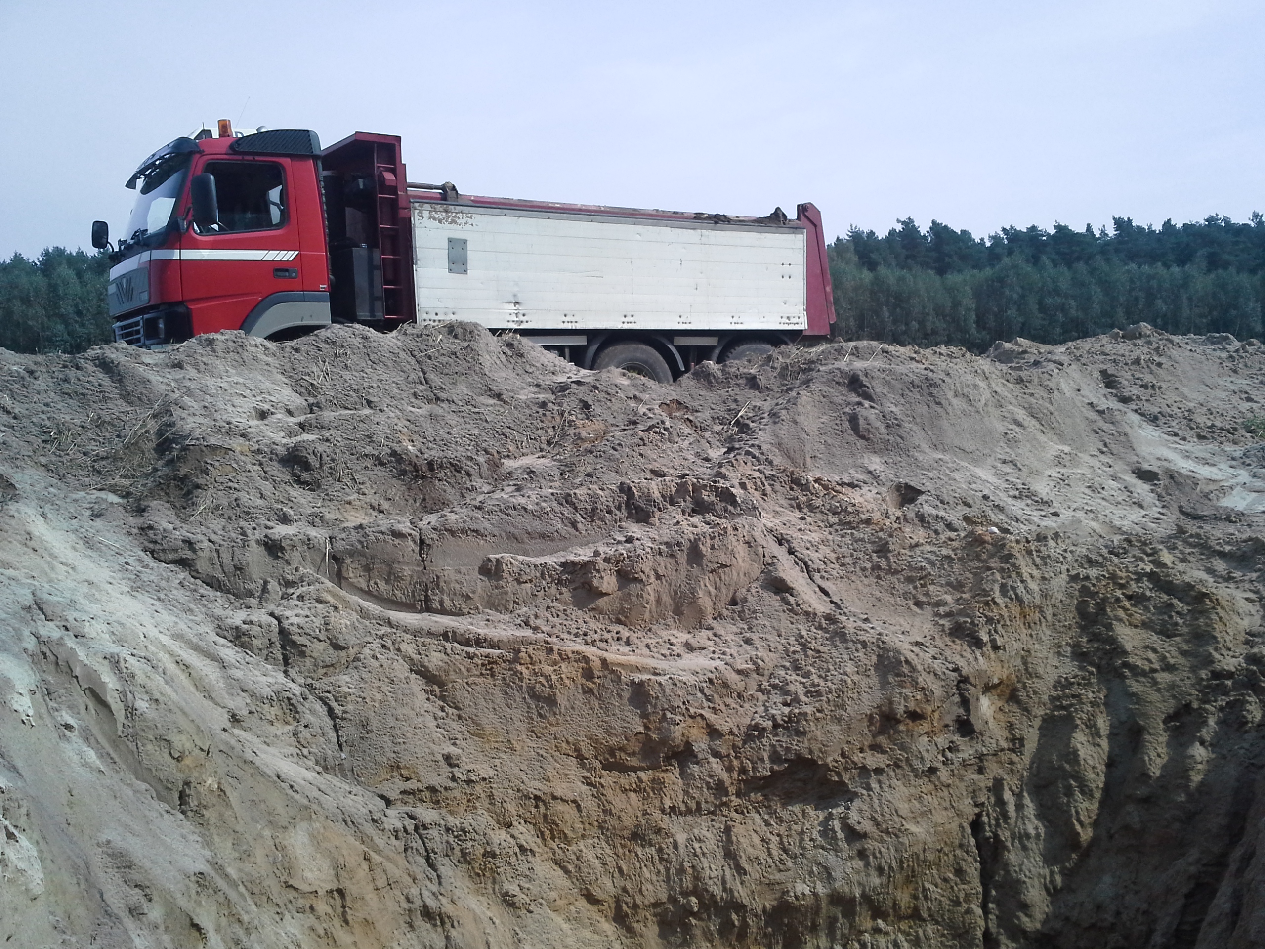 Czerwono-biała wywrotka MAN częściowo widoczna za stertą ziemi z wykopu, w tle las iglasty.