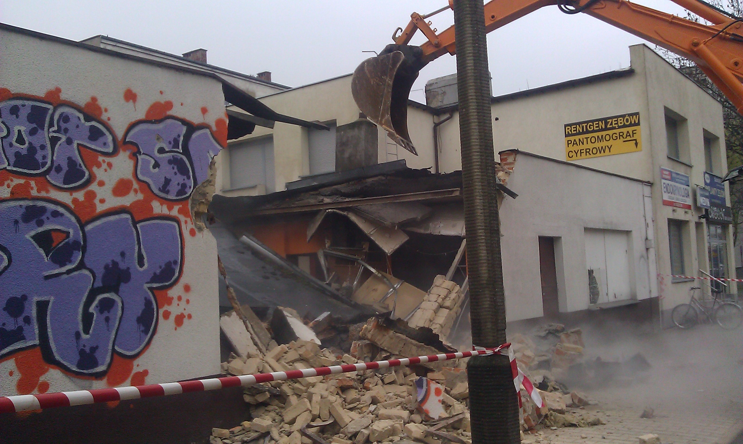 Pomarańczowa koparka burzy budynek z graffiti, gruz zabezpieczony taśmą ostrzegawczą; w tle szyld 'Rentgen Zębów'
