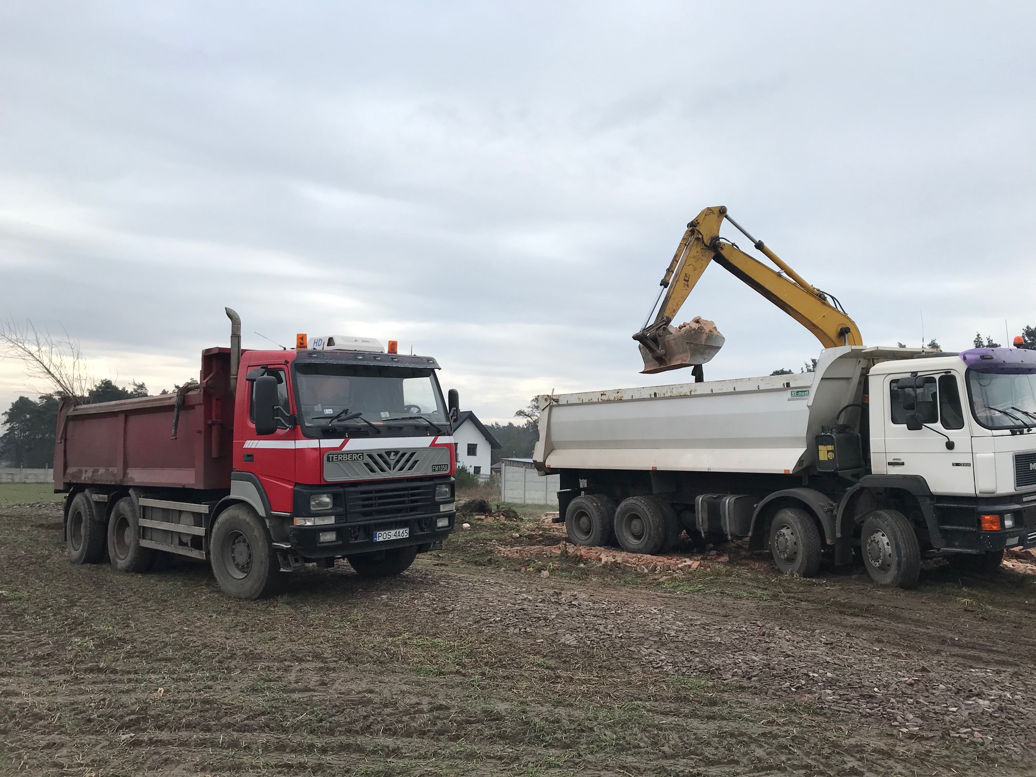 Żółta koparka załadowuje gruz do białej ciężarówki; w tle czerwona ciężarówka i dom jednorodzinny w pochmurny dzień.
