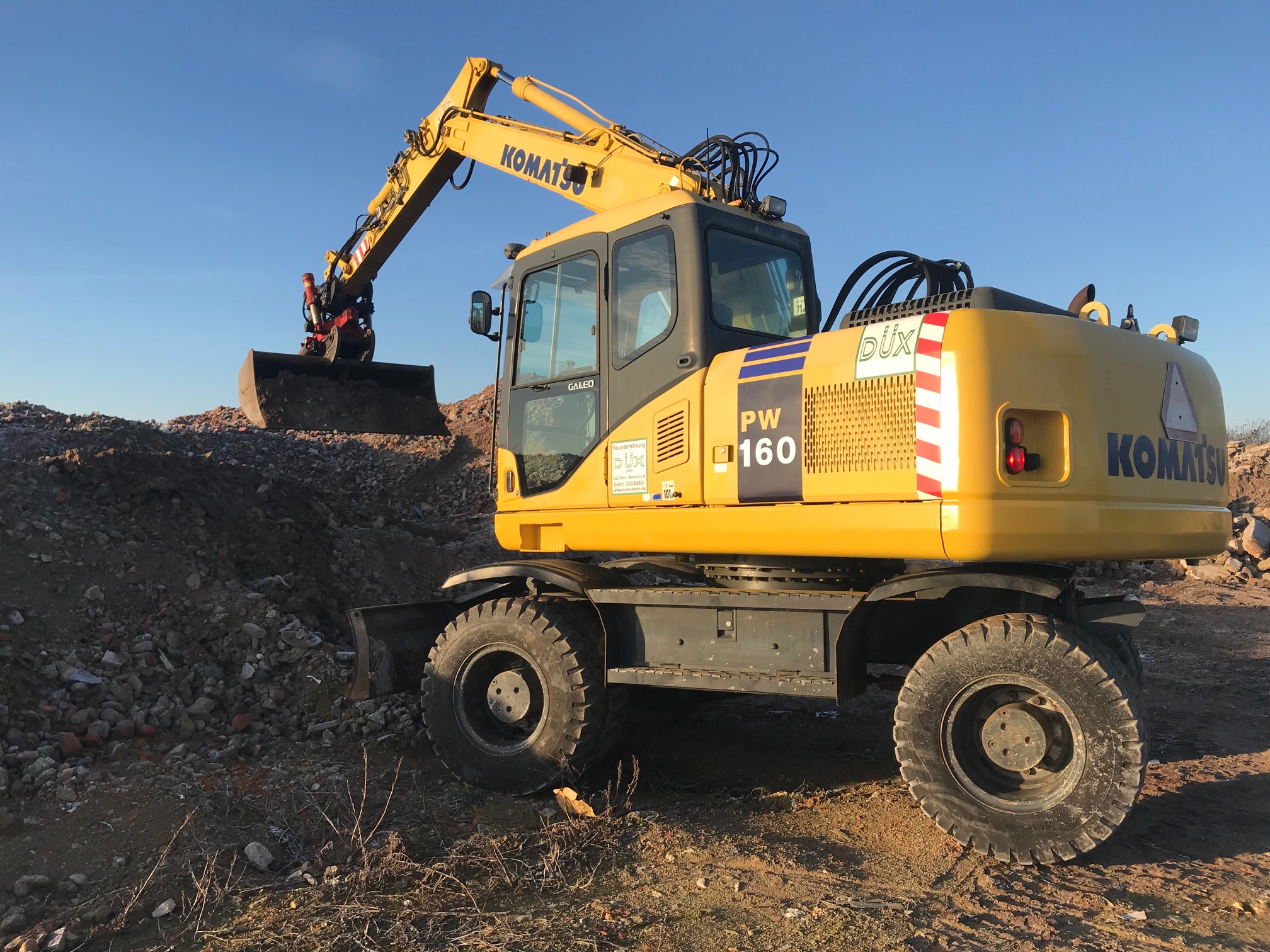 Żółta koparka kołowa Komatsu PW160 na placu budowy, z łyżką nabierającą gruz i ziemię, błękitne niebo w tle.