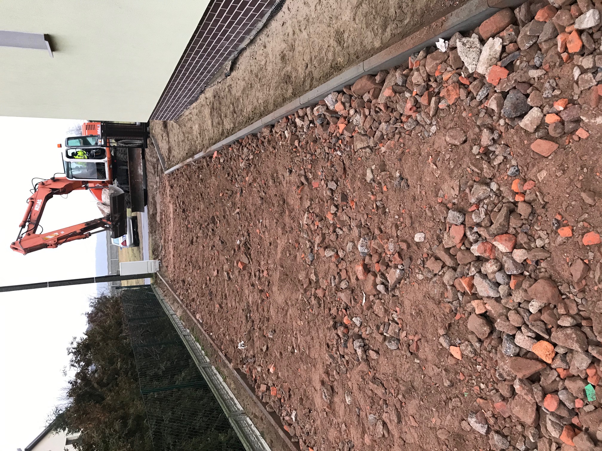 Pomarańczowa koparka Atlas przy budynku, przygotowanie terenu pod chodnik, widoczny wykop i gruz ceglany.