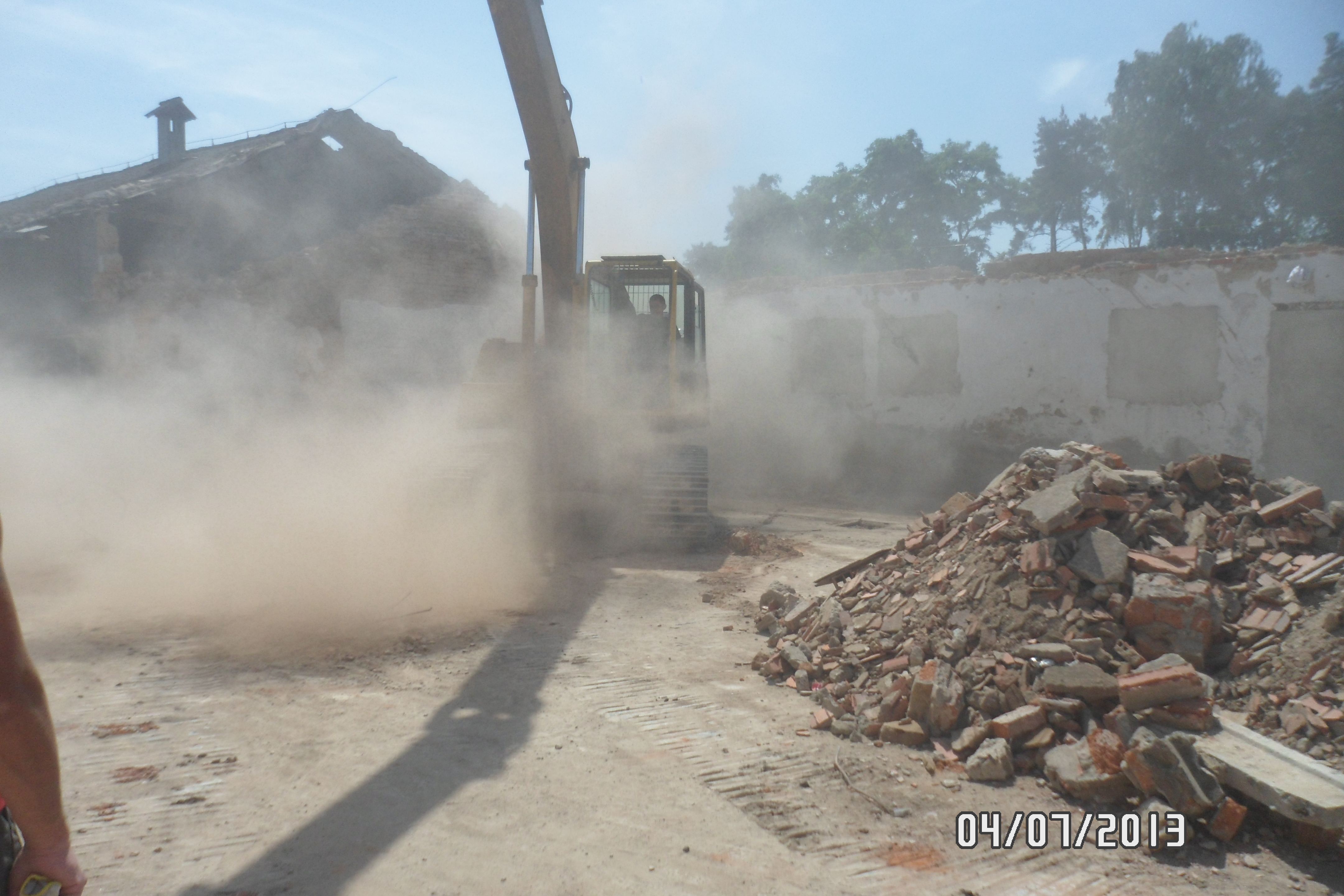 Żółta koparka w chmurze pyłu podczas rozbiórki budynku, z gruzem ceglanym w tle i datą 04/07/2013 w prawym dolnym rogu.