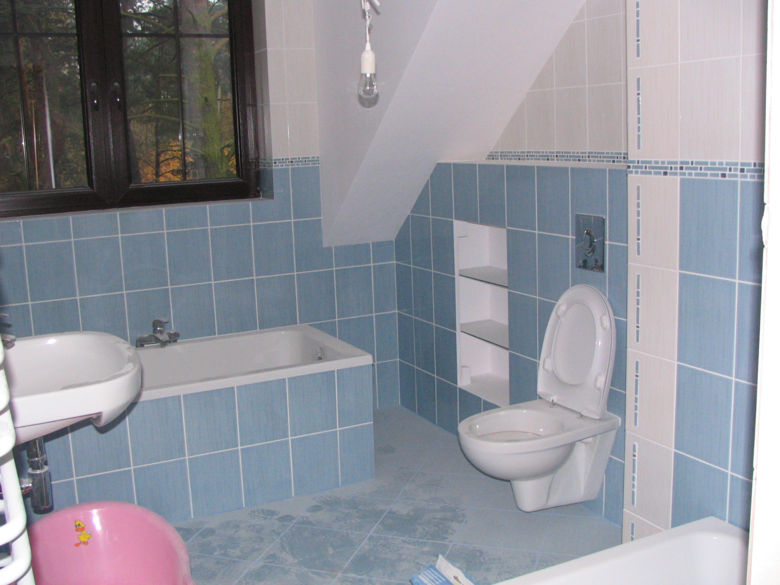 Wykończona łazienka w odcieniach błękitu i bieli, z wanną, umywalką, toaletą i wbudowanymi półkami, widok na okno z zielenią na zewnątrz.