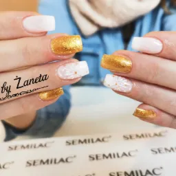 Dłonie prezentujące stylizację paznokci w Sosnowcu: jeden paznokieć biały, drugi złoty brokat, a trzeci z białym wzorem płatka śniegu i złotymi kropkami. Widoczne produkty Semilac.
