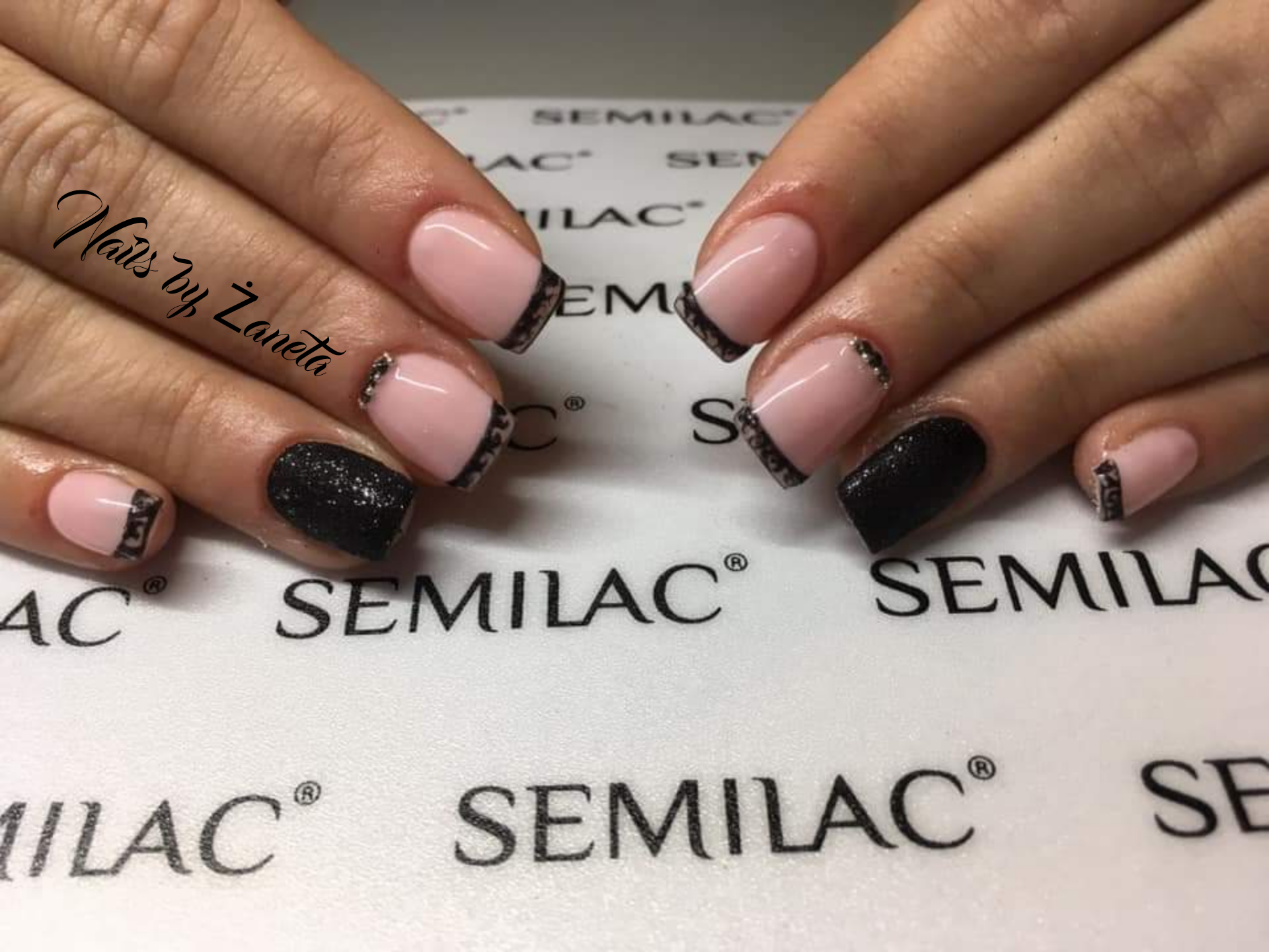 Dłonie z pomalowanymi paznokciami: jasnoróżowy lakier z czarnym wzorem na końcówkach oraz jeden paznokieć pomalowany brokatowym, czarnym lakierem, na tle podkładki marki Semilac.