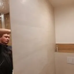 Ściana łazienki wyłożona jasnymi, beżowymi płytkami ceramicznymi o delikatnym wzorze, widoczna częściowo postać mężczyzny w odbiciu lustra, fragment kabiny prysznicowej w tle.