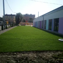 Świeżo założony trawnik z rolki przed budynkiem przedszkola z kolorowymi oknami, widoczny właz do studzienki kanalizacyjnej, w tle plac zabaw.