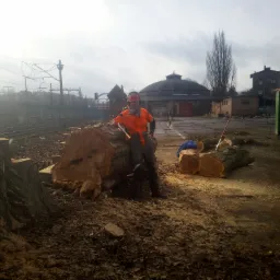 Drwal w pomarańczowej kurtce i kasku stoi obok pnia ściętego drzewa z siekierą w ręku, na tle budynków kolejowych i kontenera na odpady.