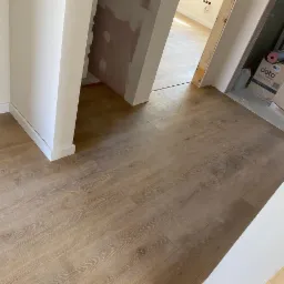 Układanie paneli podłogowych w jasnym odcieniu drewna, widoczne białe listwy przypodłogowe i fragment ściany z widocznymi śladami po szpachlowaniu.