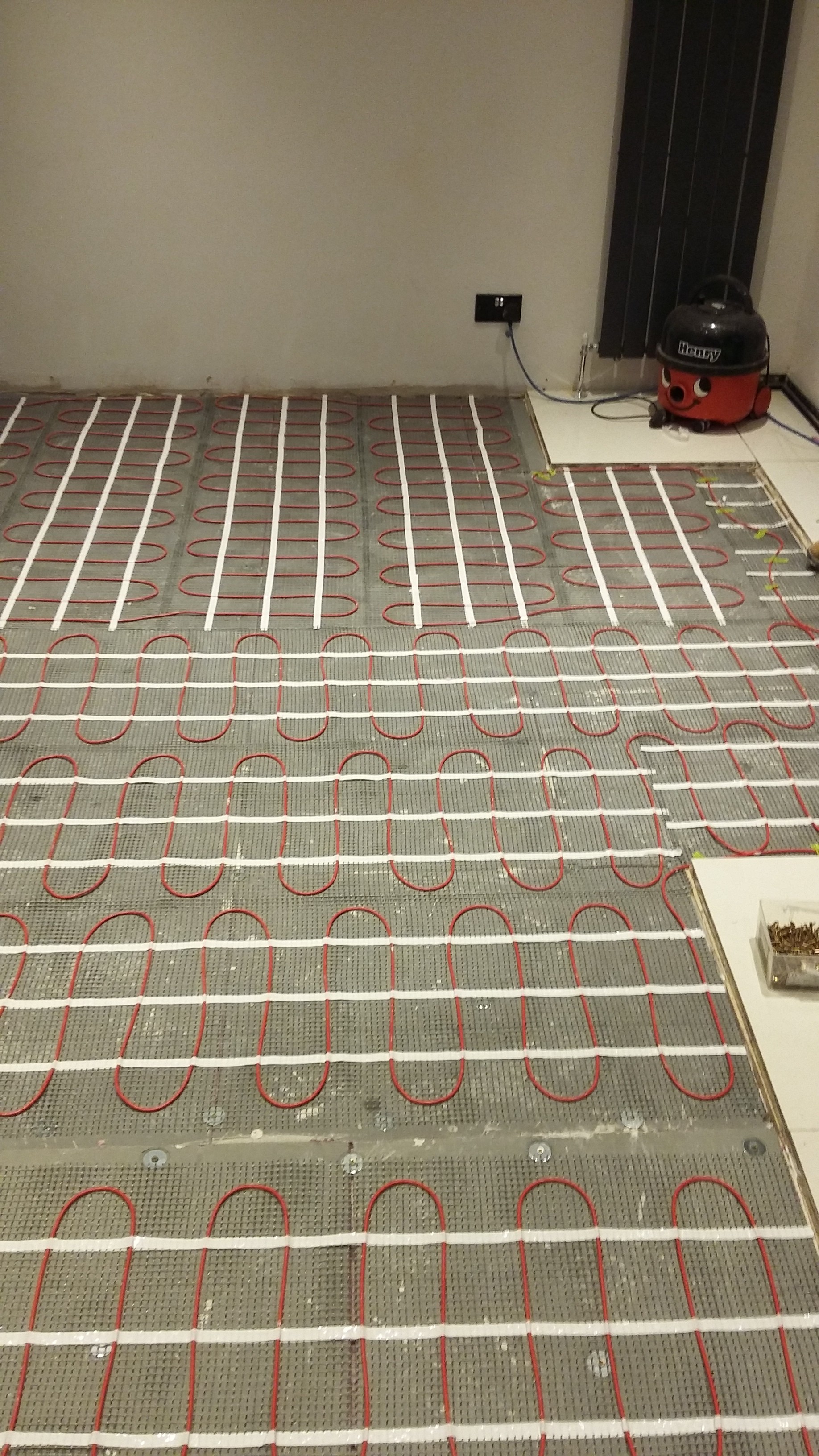Montaż ogrzewania podłogowego: czerwone kable grzewcze ułożone na szarej macie z białymi paskami mocującymi, widoczny fragment podłogi z ułożonymi płytkami i odkurzacz Henry na kafelkach.