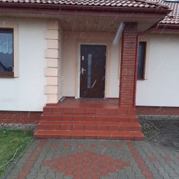 RAF-MONT - Brązowe drzwi wejściowe z srebrną klamką, widoczne z zewnątrz, z ceglanymi schodami i chodnikiem z kostki brukowej w deszczowy dzień.