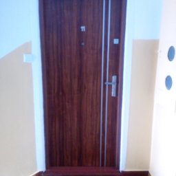 RAF-MONT - Brązowe drzwi wejściowe z numerem 11 i srebrnymi paskami, widoczne z perspektywy schodów, z wycieraczką 'Welcome Home' u dołu.