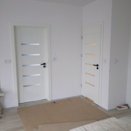 RAF-MONT - Dwa białe skrzydła drzwiowe z poziomymi, srebrnymi i złotymi wstawkami, zamontowane w nowym mieszkaniu, zabezpieczone kartonem na podłodze.