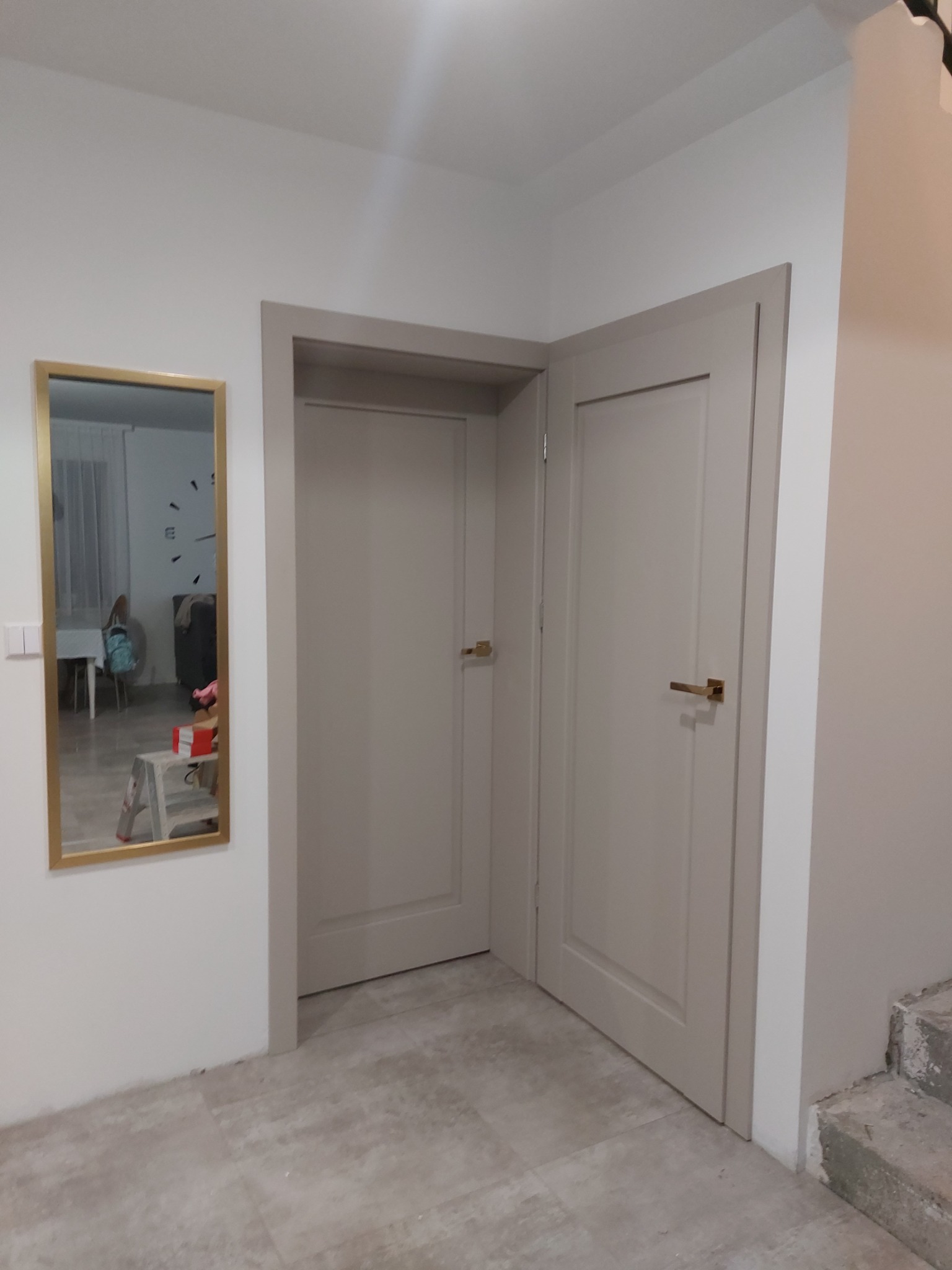 Dwa beżowe skrzydła drzwiowe z złotymi klamkami w rogu pokoju, widok na lustro w złotej ramie odbijające fragment pokoju z zegarem na ścianie.