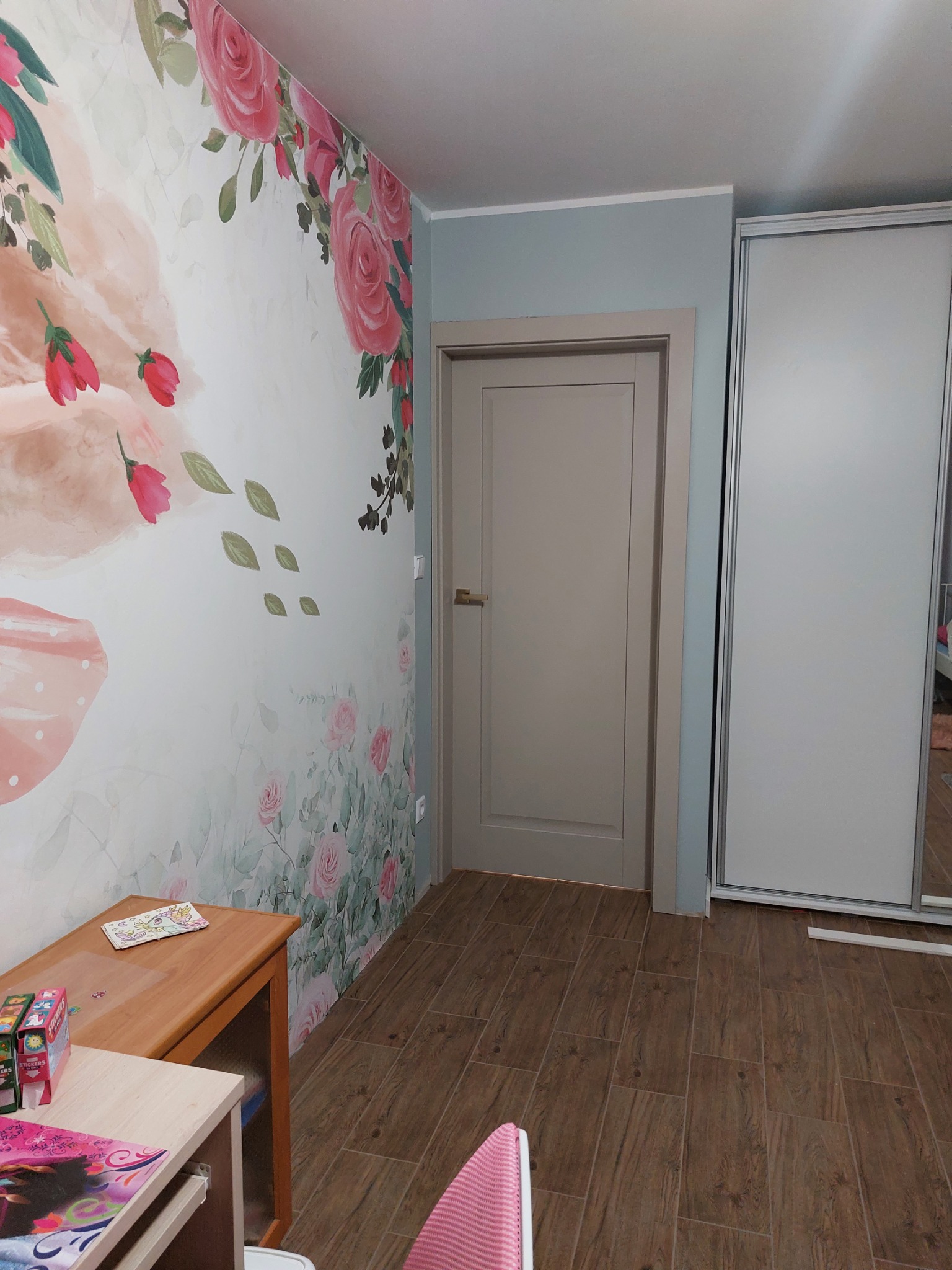 Jasnoszare drzwi wewnętrzne z ościeżnicą w pokoju dziecięcym z fototapetą w kwiaty i brązowymi panelami podłogowymi imitującymi drewno, obok biała szafa przesuwna z lustrem.