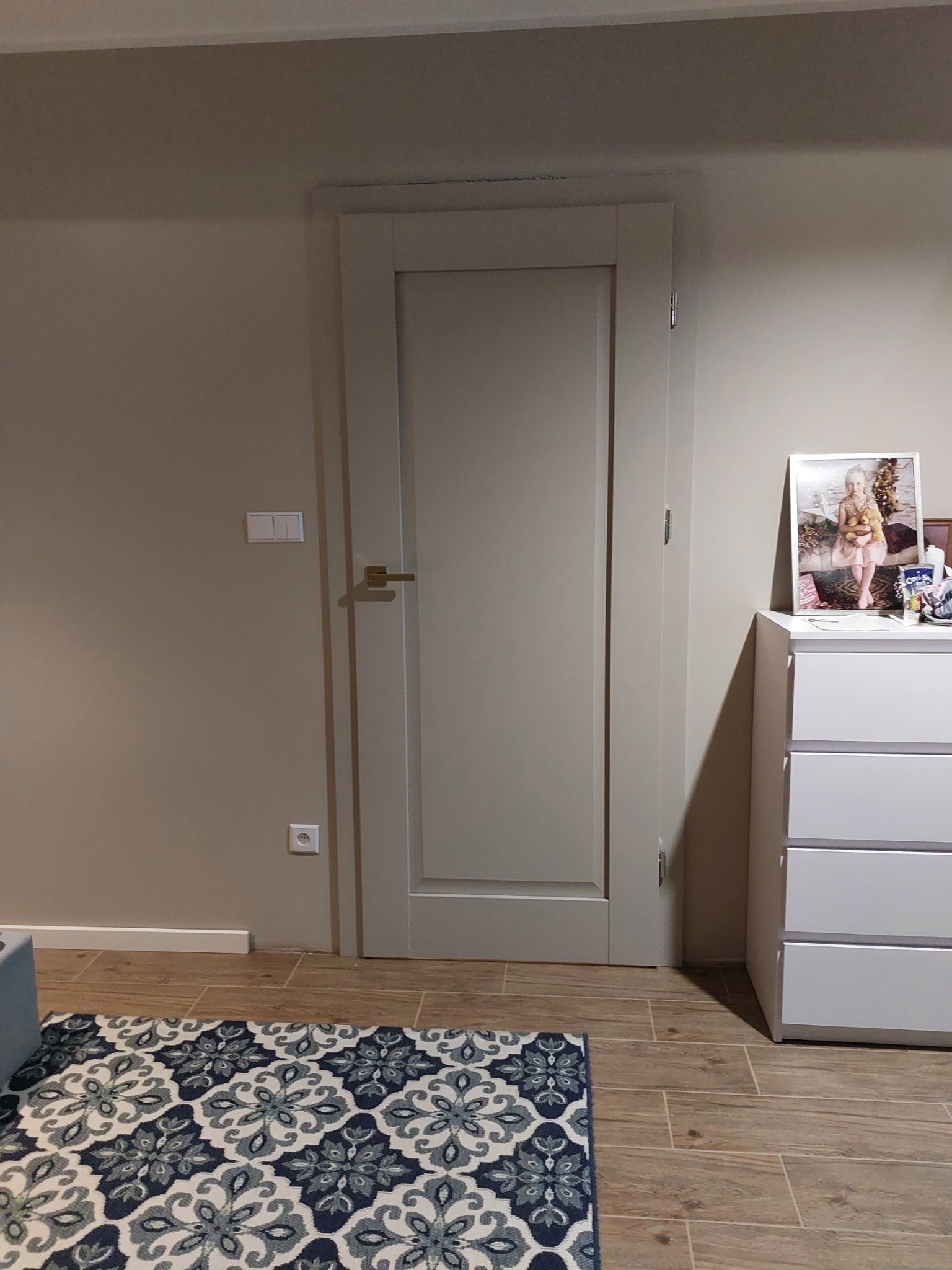 Zamknięte, białe drzwi wewnętrzne z prostą klamką na tle beżowej ściany i drewnopodobnej podłogi, fragment wzorzystego dywanu.