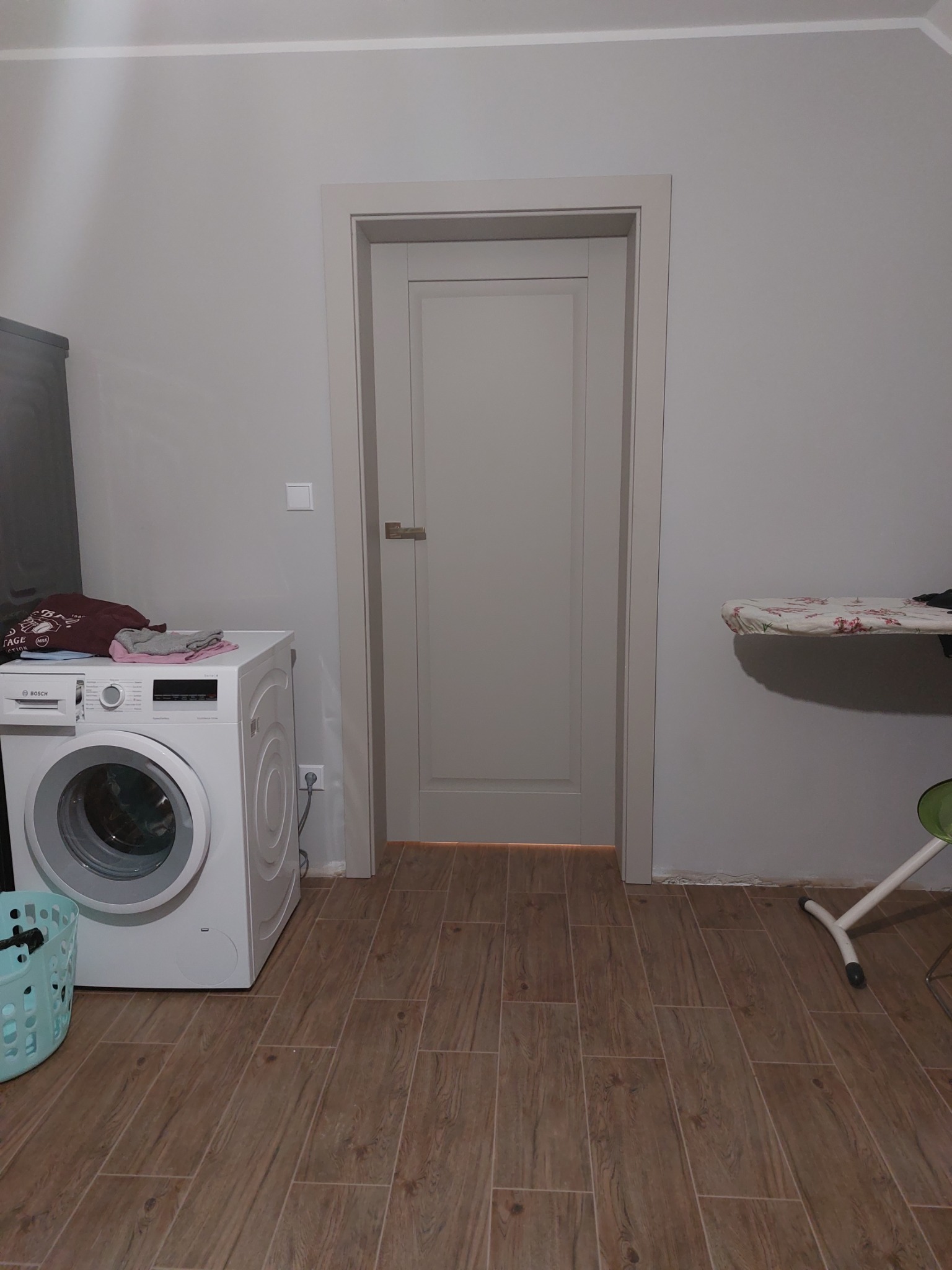 Szara, zamknięta płycinowa, wewnętrzna skrzydło drzwiowe z srebrną klamką, widoczne na tle płytek podłogowych imitujących drewno, pralka, deska do prasowania i kosz na pranie w pomieszczeniu.
