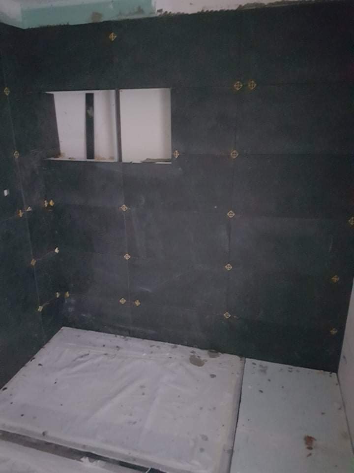 Ciemne płytki glazurnicze z żółtymi krzyżykami dystansowymi na ścianie łazienki w trakcie remontu, widoczna wnęka w ścianie i zabezpieczona folią podłoga.