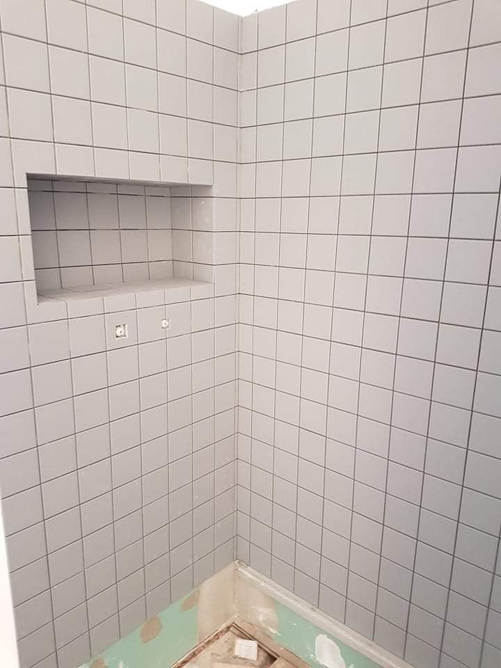 Świeżo położone, kwadratowe, szare płytki na ścianach łazienki z wnęką, widoczne przygotowania do dalszych prac wykończeniowych przy podłodze.