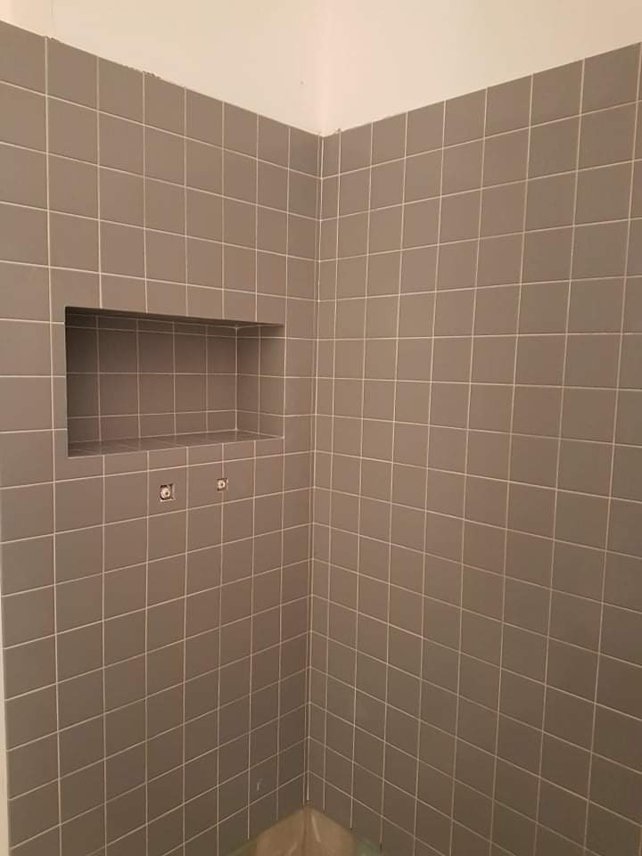 Ściana wyłożona szarymi, kwadratowymi płytkami z wnęką oraz widocznymi otworami na armaturę.