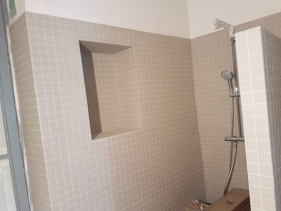 Ściana łazienki wyłożona jasnymi, kwadratowymi płytkami z wnęką, widoczny panel prysznicowy z folią ochronną na głowicy.