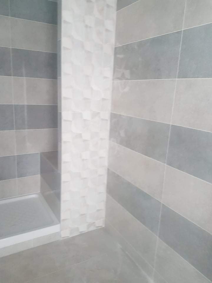 Wykończona łazienka z kabiną prysznicową, ściany wyłożone szarymi i beżowymi pasami płytek oraz białą ścianą z trójwymiarowym wzorem.