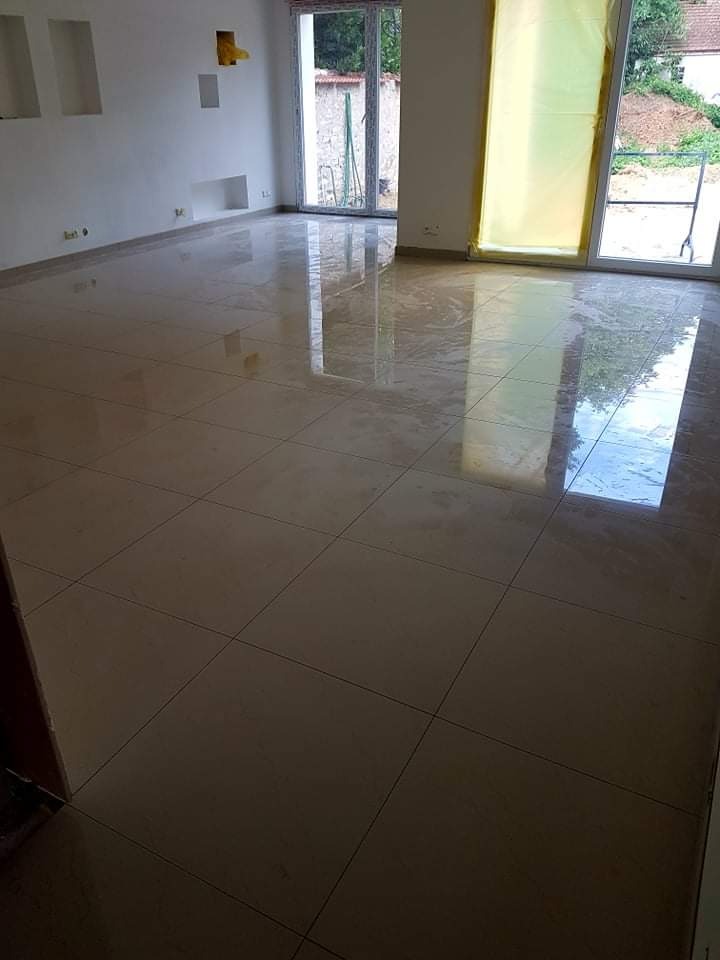 Wykończone wnętrze z błyszczącą, beżową podłogą z płytek ceramicznych, duże okna z widokiem na zieleń za oknem, ściany w kolorze złamanej bieli, wnęki w ścianie. Jedno z okien zabezpieczone folią...