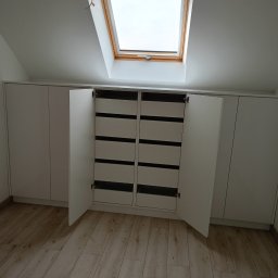 Mebstar - Biała szafa na poddaszu ze skośnym oknem dachowym. Otwarta szafa z szufladami, jasna podłoga. Minimalistyczny design, zabudowa pod skosem.